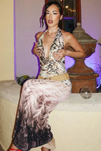 Woman wearing a sleeveless, halter-neck, abstract print maxi dress with a plunging neckline, sitting on a stone ledge.
