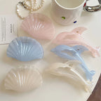 Collection of pastel colored dolphin and seashell hair claw clips and a pearl necklace on a white surface