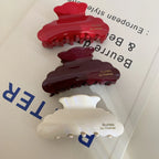 A top-down view of three glossy hair claw clips in red, burgundy, and cream, stacked on a surface with text. The clips are cloud-shaped with embossed gold lettering.