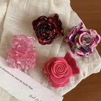 Four rose-shaped hair claw clips in various colors including pink, purple, and red tortoise shell pattern, arranged on a white cloth with a piece of paper