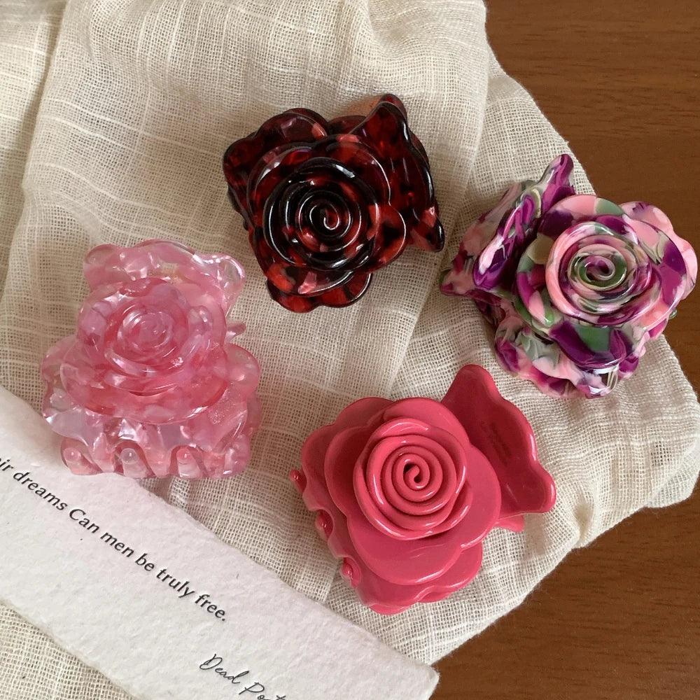 Four rose-shaped hair claw clips in various colors including pink, purple, and red tortoise shell pattern, arranged on a white cloth with a piece of paper