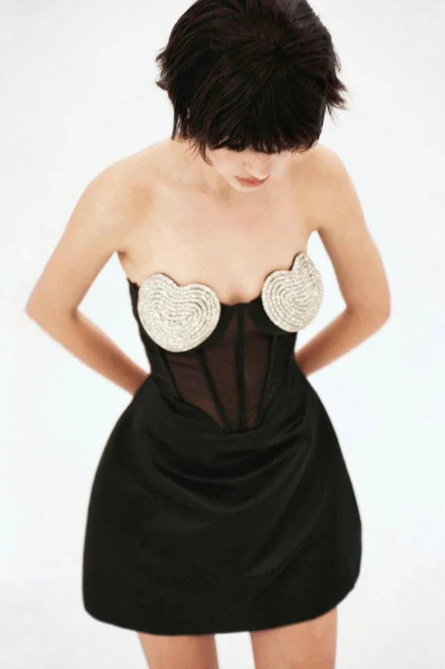Model looking down while wearing a black strapless dress with heart-shaped embellishments on the bust and a corset bodice.