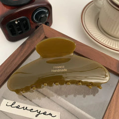 A single olive green hair claw clip, made of acetate, with 'FRANCE Handmade' printed in gold, resting on a wooden frame with a sheer fabric underneath.