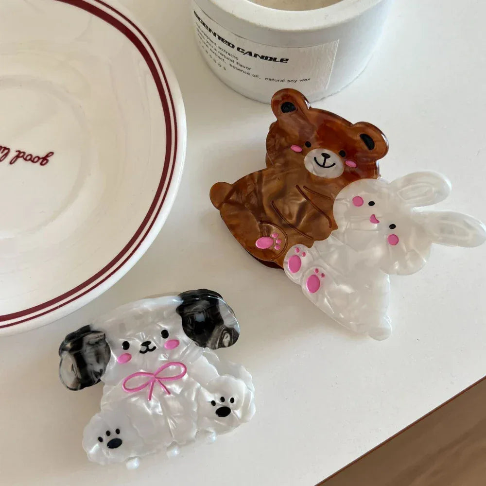 A white bunny and a brown bear hair claw clip are shown next to a white dog hair claw clip on a white surface, with a candle and a book in the background.