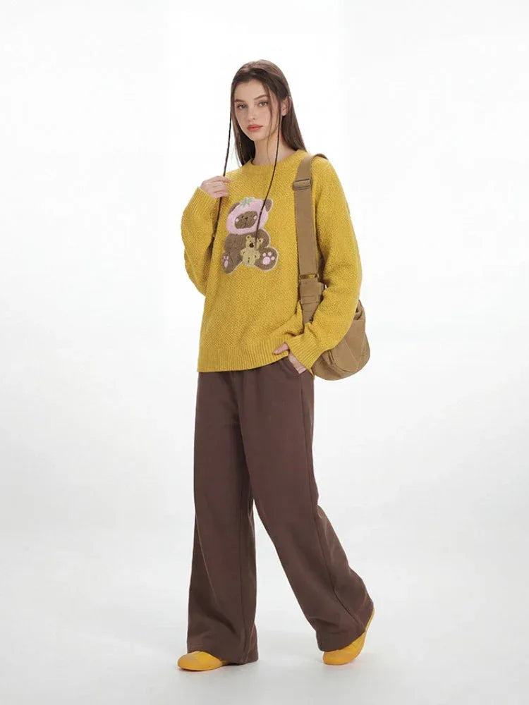 Woman in a yellow crew neck sweater with a bear graphic and brown wide-leg pants, standing with one hand in her pocket. Secondary image