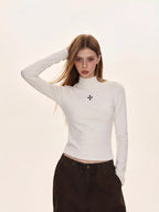 Woman wearing a white turtleneck top with a black embroidered cross, paired with dark brown cargo pants.