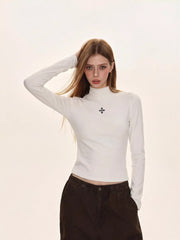 Woman wearing a white turtleneck top with a black embroidered cross, paired with dark brown cargo pants.