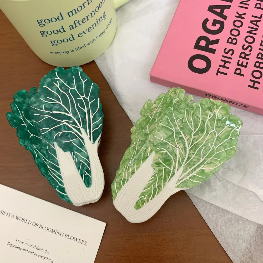 Two napa cabbage-shaped accessories, one dark green and one light green, are displayed on a wooden surface next to a pink book and a white mug.