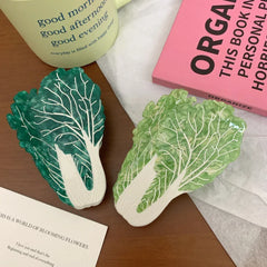 Two napa cabbage-shaped accessories, one dark green and one light green, are displayed on a wooden surface next to a pink book and a white mug.