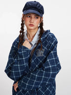 Front detail of a model wearing a blue plaid hooded jacket with an embroidered logo, light blue collared shirt, and a navy blue cap.