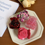 Four rose-shaped hair claw clips in various colors including pink, purple, and red tortoise shell pattern, arranged on a white plate with a book and yellow flowers