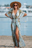 Woman wearing a flowing, tie-waist maxi dress with a watercolor-inspired print and a straw hat, standing on a beach with the ocean in the background.