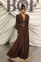 A woman in a brown draped maxi dress with a cinched waist and dramatic sleeves stands with her hand on her hip.
