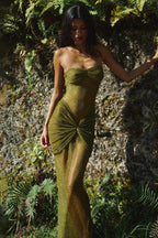 Woman wearing a strapless, shimmering olive green gown with ruched detailing and a knotted front accent, posing outdoors in a natural setting.