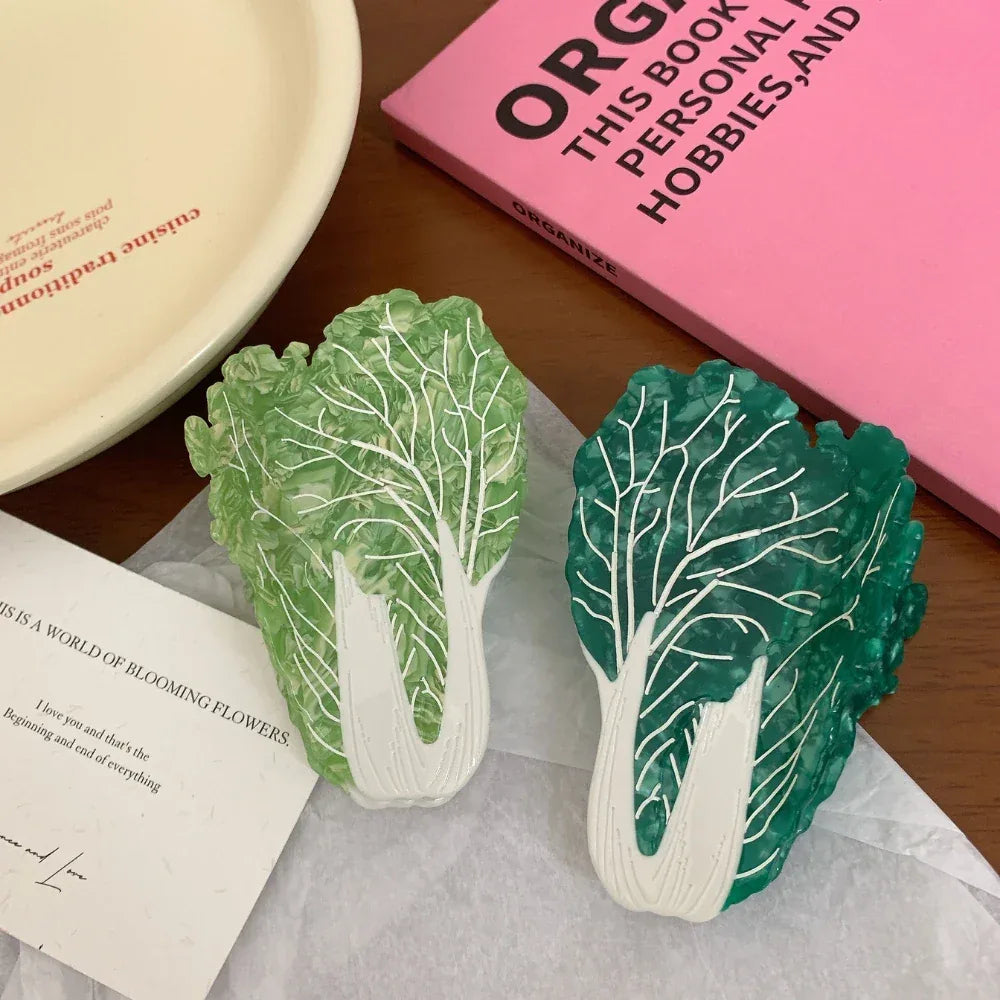 Two napa cabbage-shaped accessories, one dark green and one light green, are arranged on a wooden surface with a pink book and a greeting card.