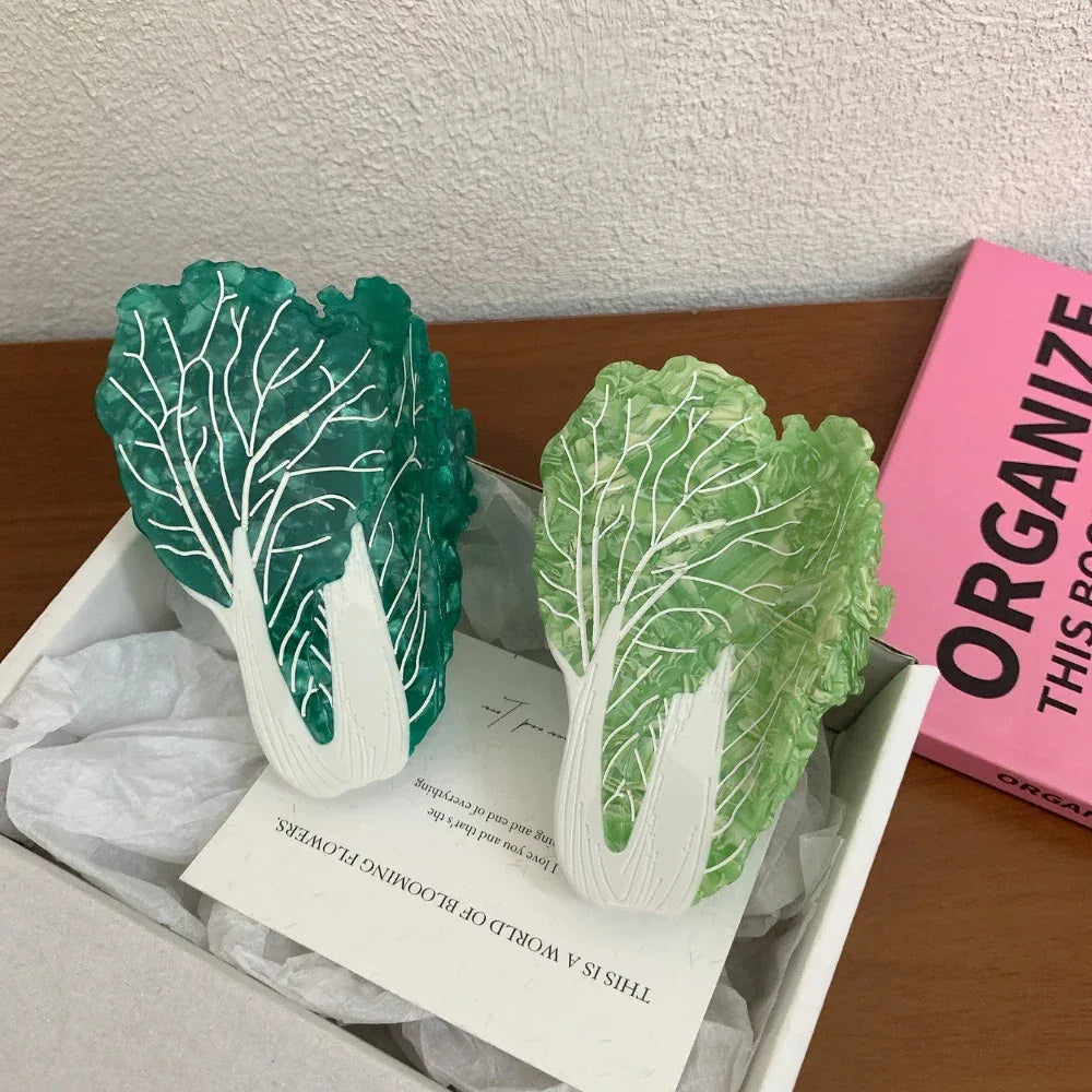 Two napa cabbage-shaped accessories, one dark green and one light green, are presented in a white box with a greeting card.