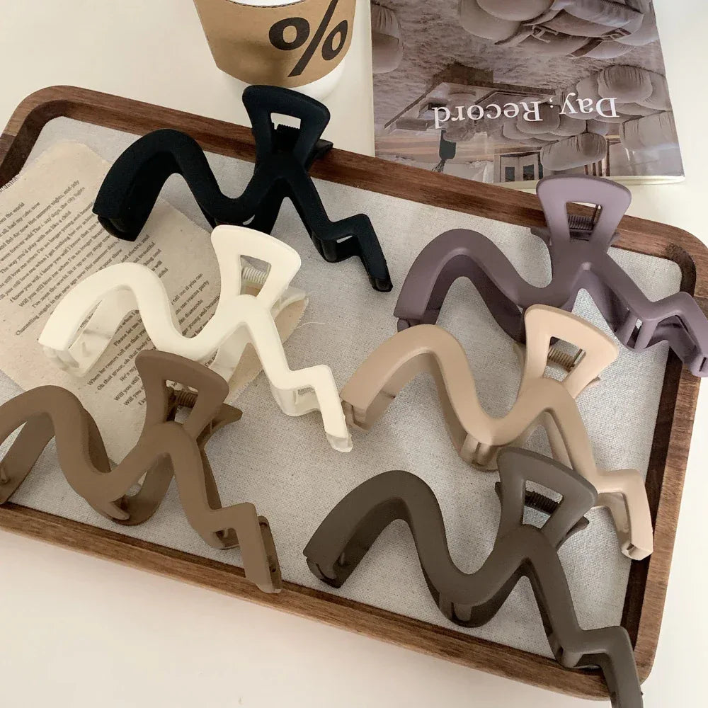 A collection of wavy claw clips in various colors including black, white, purple, beige, and brown, arranged on a wooden tray.