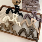 A collection of wavy claw clips in various colors including black, white, purple, beige, and brown, arranged on a wooden tray.