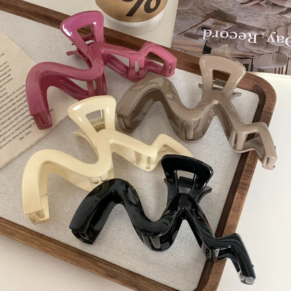 A group of five wavy claw clips in pink, beige, cream, black, and dark brown, arranged on a linen-like surface within a wooden tray.
