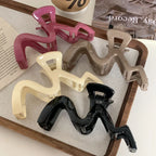 A group of five wavy claw clips in pink, beige, cream, black, and dark brown, arranged on a linen-like surface within a wooden tray.