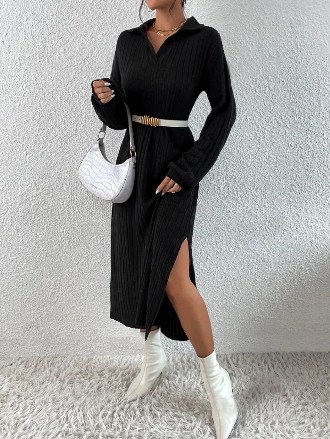 Woman wearing a black long-sleeved ribbed knit dress with a high slit, white boots, a white handbag, and a white belt with gold accents.