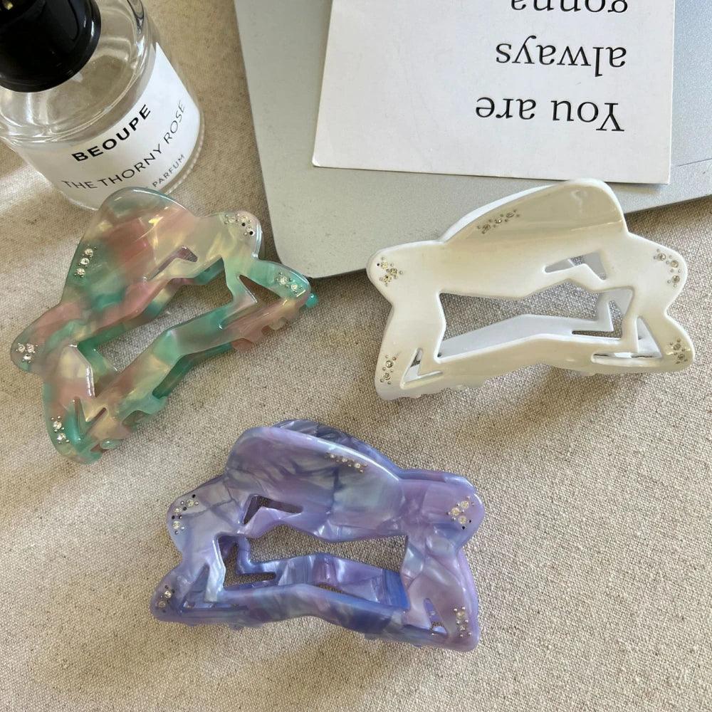 three abstract figure hair claw clips in marbled green/pink/brown, white, and purple arranged on a textured surface