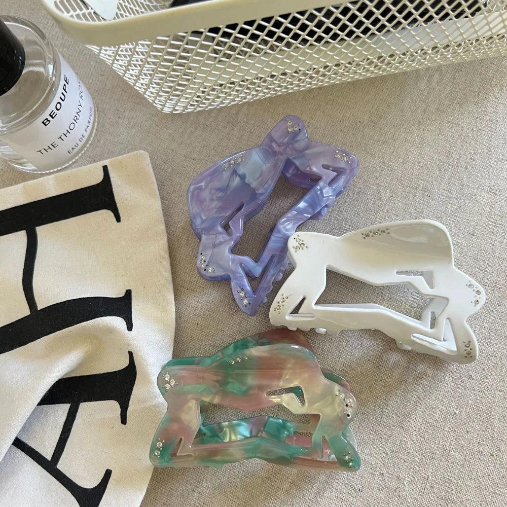 overhead view of three abstract figure hair claw clips in purple, white, and marbled green/pink/brown on a textured surface with a bottle of perfume and a white basket