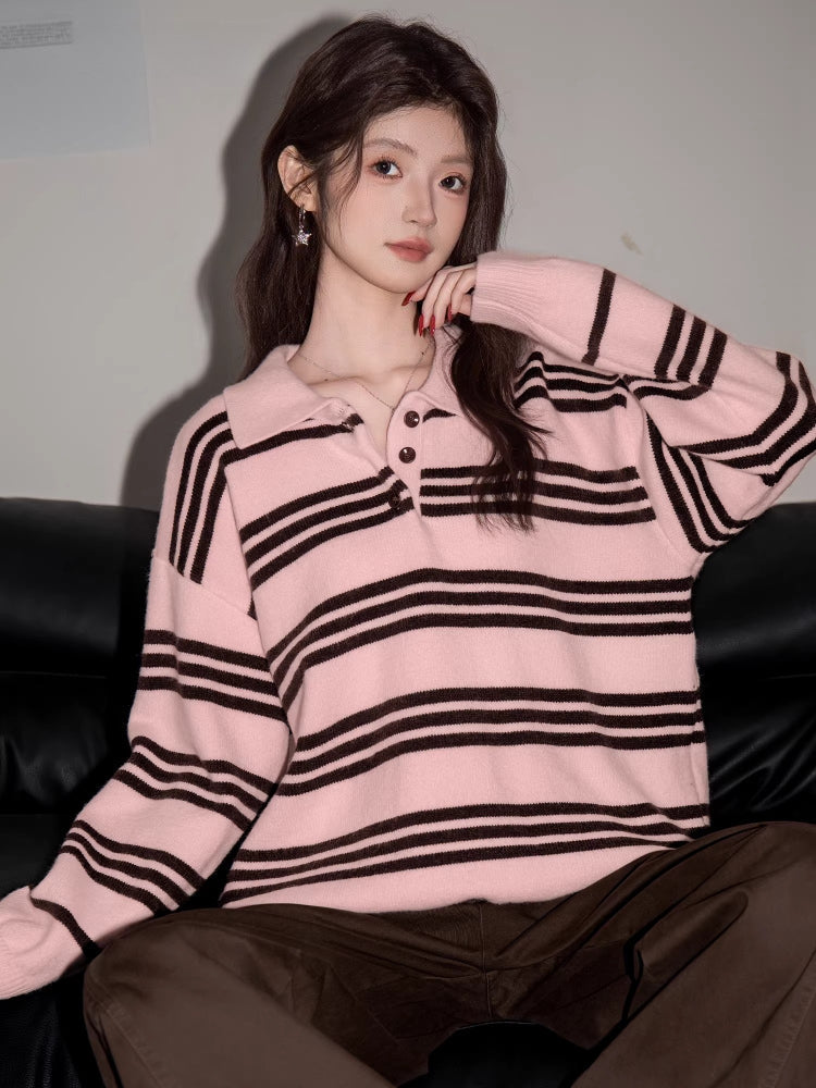 Woman wearing a pink and black striped knit polo sweater and brown pants, sitting on a black couch, posing with her hand on her chin.