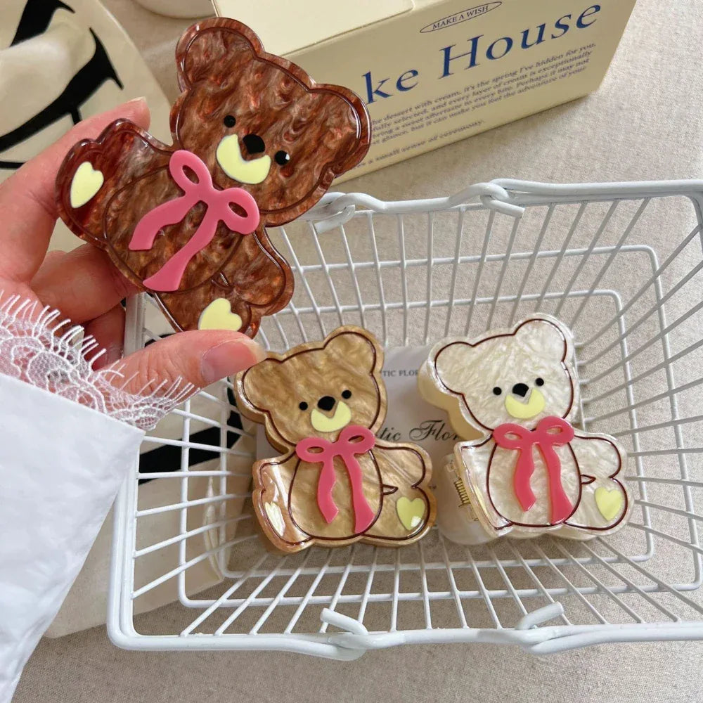 A hand holding a brown teddy bear hair clip, with two other teddy bear clips in a white basket below.