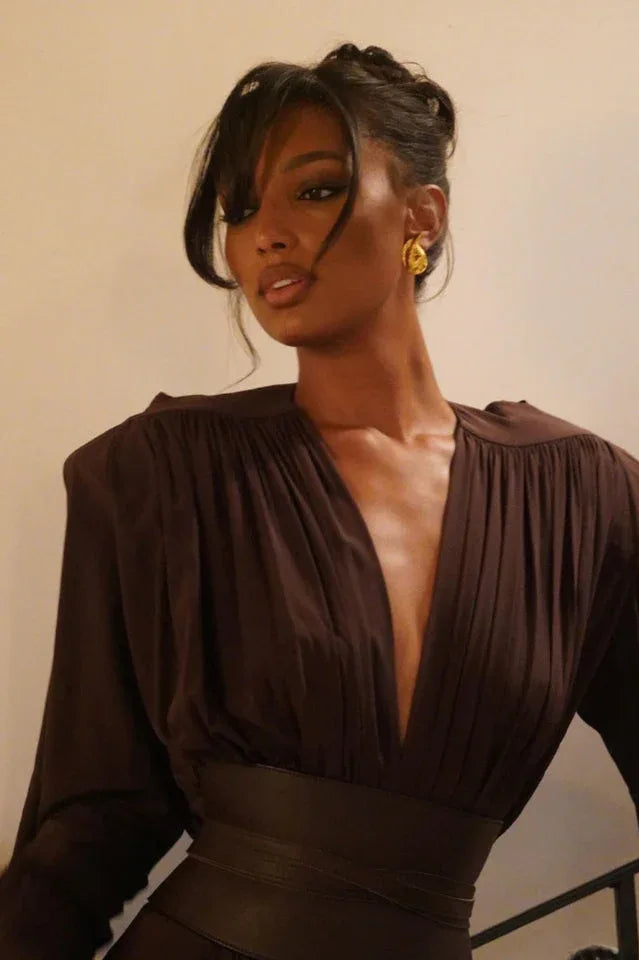 Close-up of a model's face and upper body, showcasing the neckline and earrings of a brown dress.
