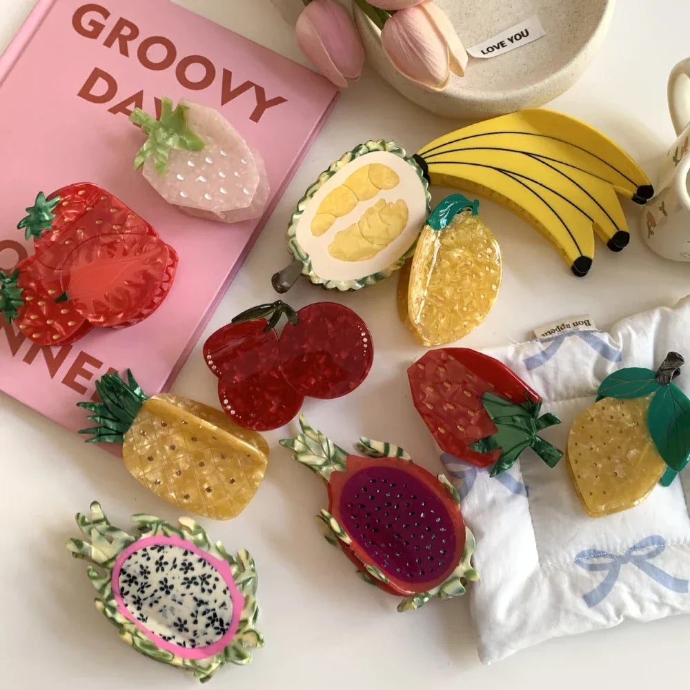 An overhead view of a variety of fruit-shaped hair clips, such as bananas, strawberries, cherries, dragon fruit, durian, pineapple, and lemons, displayed with other decorative items.