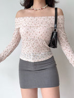 Woman wearing a pink floral off-the-shoulder long-sleeved top, pearl and rose necklace, and a black patent handbag with gray mini skirt.