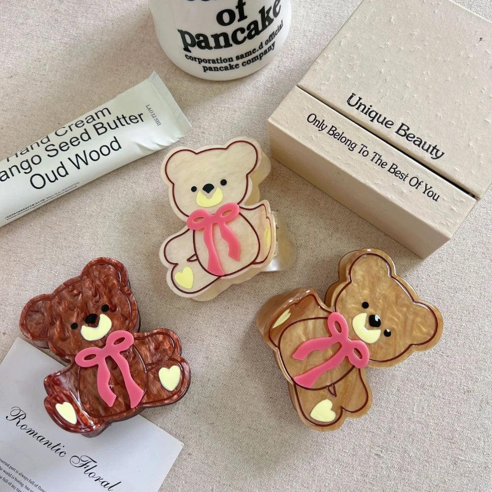 Three teddy bear hair clips in brown, beige, and cream colors, arranged with a tube of hand cream and a box.