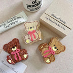 Three teddy bear hair clips in brown, beige, and cream colors, arranged with a tube of hand cream and a box.
