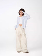 Woman posing with hand on forehead, wearing a light blue cropped knit cardigan, white crop top, and cream wide-leg pants.