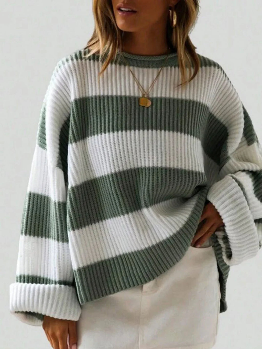 Woman wearing a green and white striped knit sweater with rolled-up sleeves and a gold pendant necklace, paired with white bottoms.