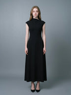 Woman in a black mock neck cap sleeve A-line dress standing against a grey background.
