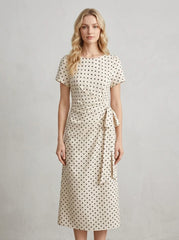 Woman wearing a cream and black polka dot midi wrap dress with short sleeves, standing against a white textured wall.