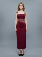 Woman wearing a long, burgundy slip dress with lace waist detailing, standing against a gray background.