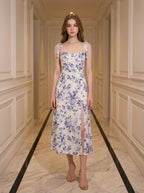 Woman wearing a floral print midi dress with tie shoulder straps and a high slit, standing in a hallway.