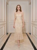 Woman wearing a cream-colored sleeveless midi dress with floral embellishments and a ruffled hem, standing in a hallway.