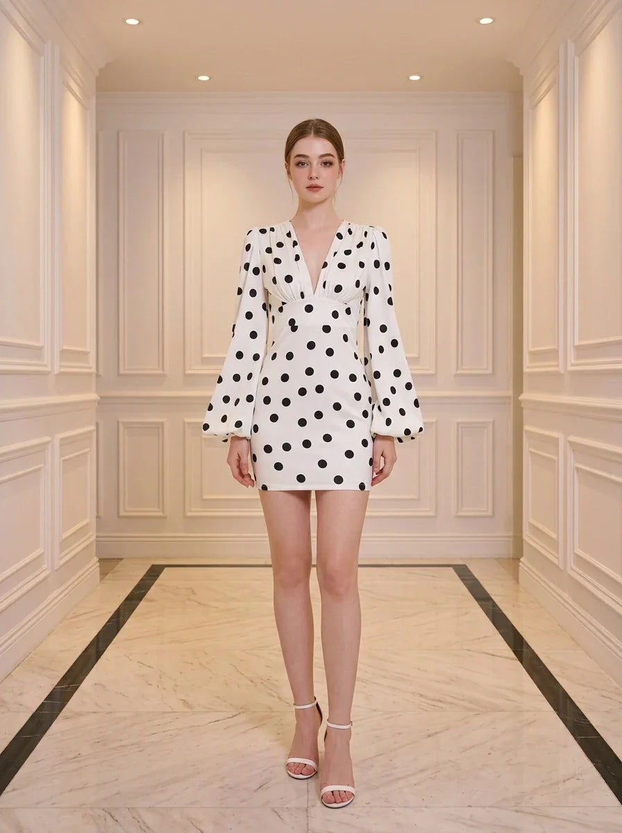 Woman standing in a white hallway wearing a white polka dot mini dress with long bishop sleeves and white heels.
