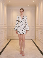 Woman standing in a white hallway wearing a white polka dot mini dress with long bishop sleeves and white heels.