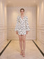 Woman standing in a white hallway wearing a white polka dot mini dress with long bishop sleeves and white heels.