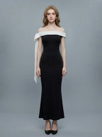 Woman wearing a black off-the-shoulder mermaid gown with a white sash and bow detail, standing against a grey background.