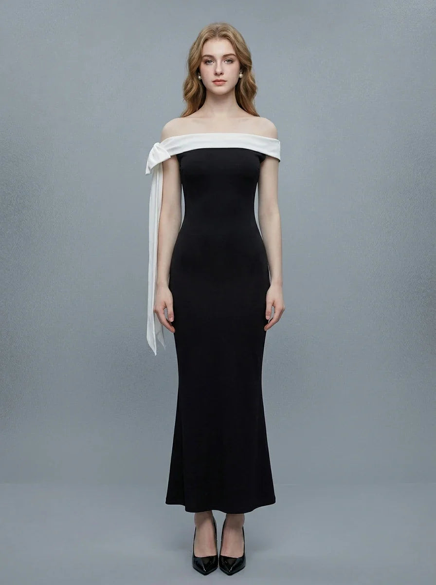 Woman wearing a black off-the-shoulder mermaid gown with a white sash and bow detail, standing against a grey background.