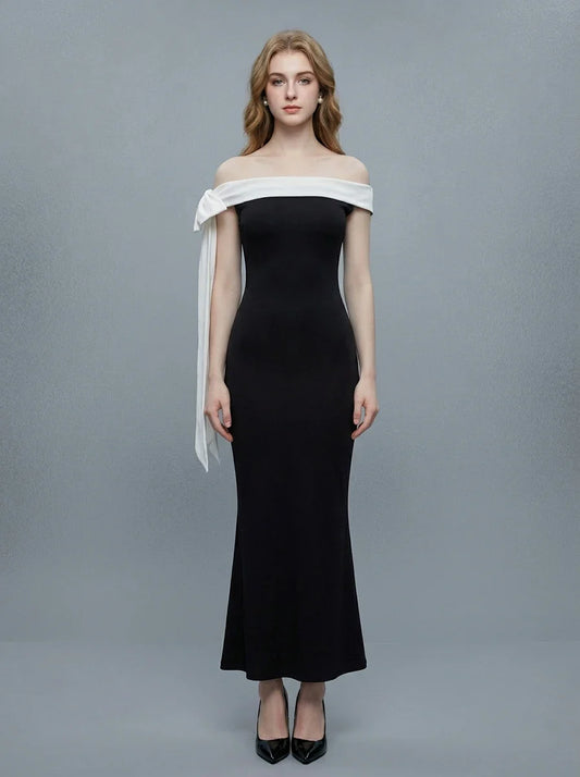 Woman wearing a black off-the-shoulder mermaid gown with a white sash and bow detail, standing against a grey background.