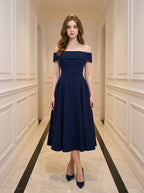 Woman wearing a navy blue off-the-shoulder midi dress with a pleated skirt, standing in a hallway.