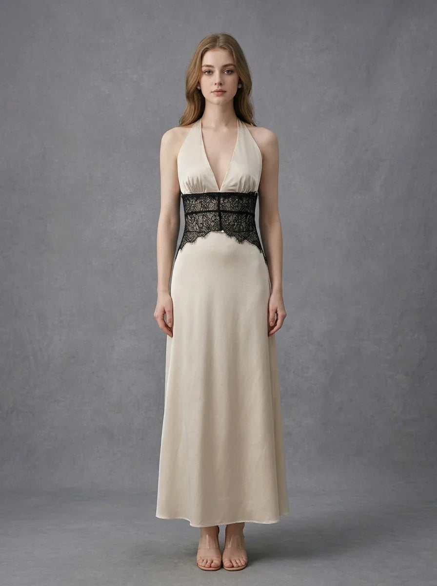 Woman in a cream satin maxi dress with black lace waist detail, standing against a grey backdrop.