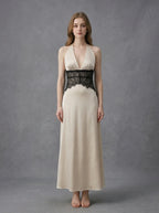 Woman in a cream satin maxi dress with black lace waist detail, standing against a grey backdrop.